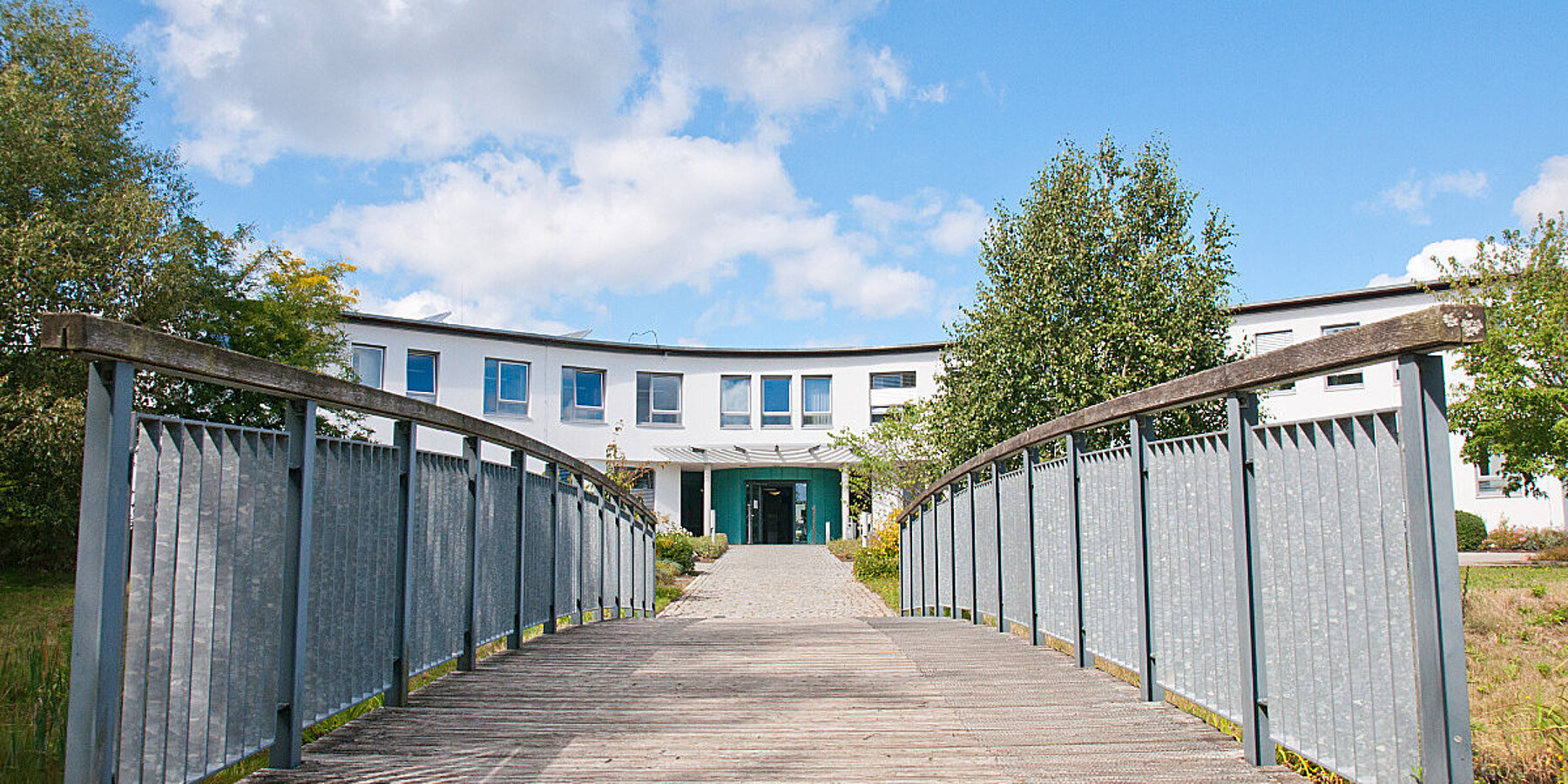 Panorama der Holzbrücke vor dem Haupteingang zur Psychiatrischen Klinik Uelzen, darüber blauer Himmel mit weißen Wolken