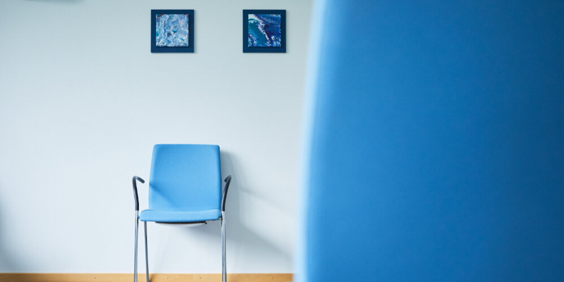 Links blauer Stuhl vor weißer Wand mit zwei kleinen blauen Wandbildern, vorne rechte Bildhälfte unscharfe blaue Stuhllehne