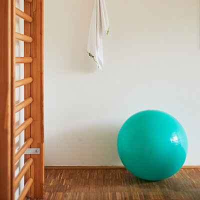 Bild eines Therapieraums: Links eine Sprossenwand, daneben ein großer Gymnastikball, an der Wand hängt ein Handtuch an einem Haken.